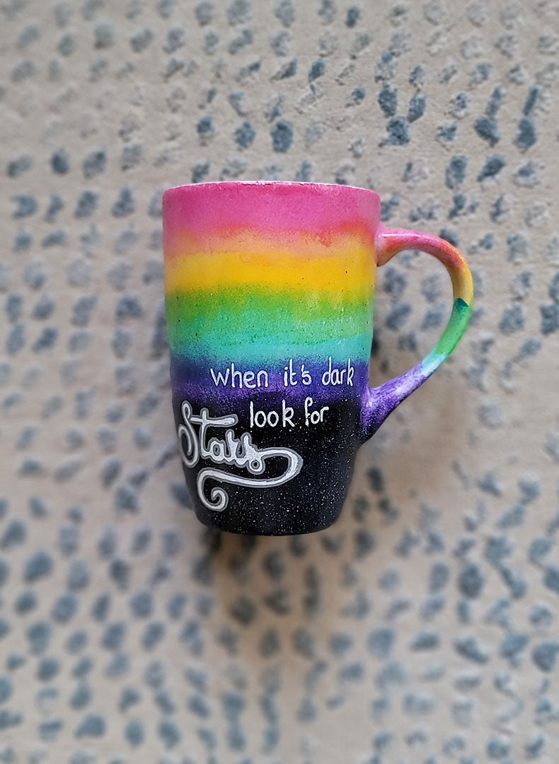 A Mug with the top half painted in rainbow and the bottom in dark galaxy, with the words on the dark galaxy "when its dark look for stars"