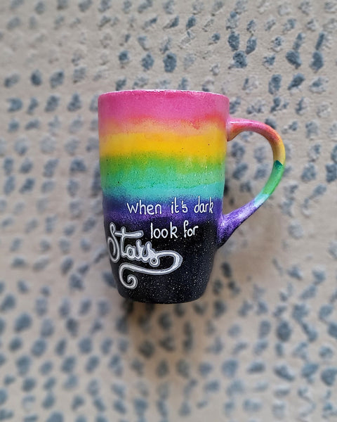 A Mug with the top half painted in rainbow and the bottom in dark galaxy, with the words on the dark galaxy "when its dark look for stars"