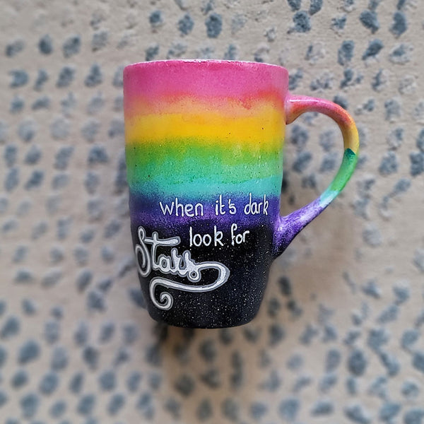 A Mug with the top half painted in rainbow and the bottom in dark galaxy, with the words on the dark galaxy "when its dark look for stars"