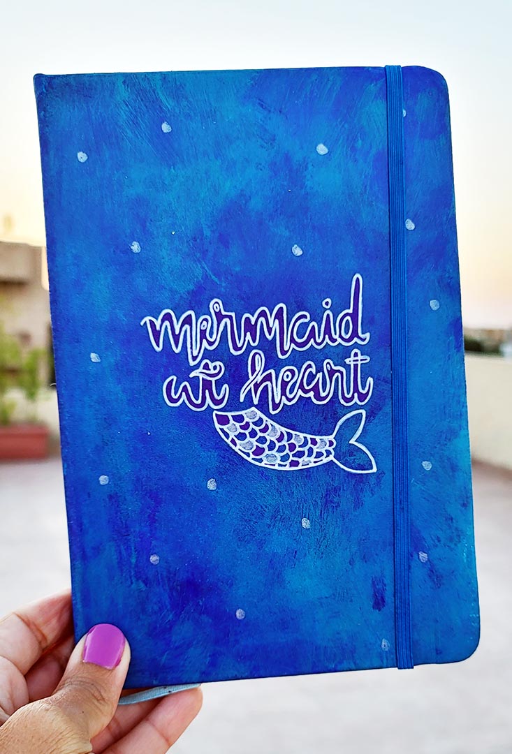 A blue hand painted Notebook with the words "Mermaid at heart" with a mermaid tail painted below it