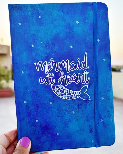 A blue hand painted Notebook with the words "Mermaid at heart" with a mermaid tail painted below it