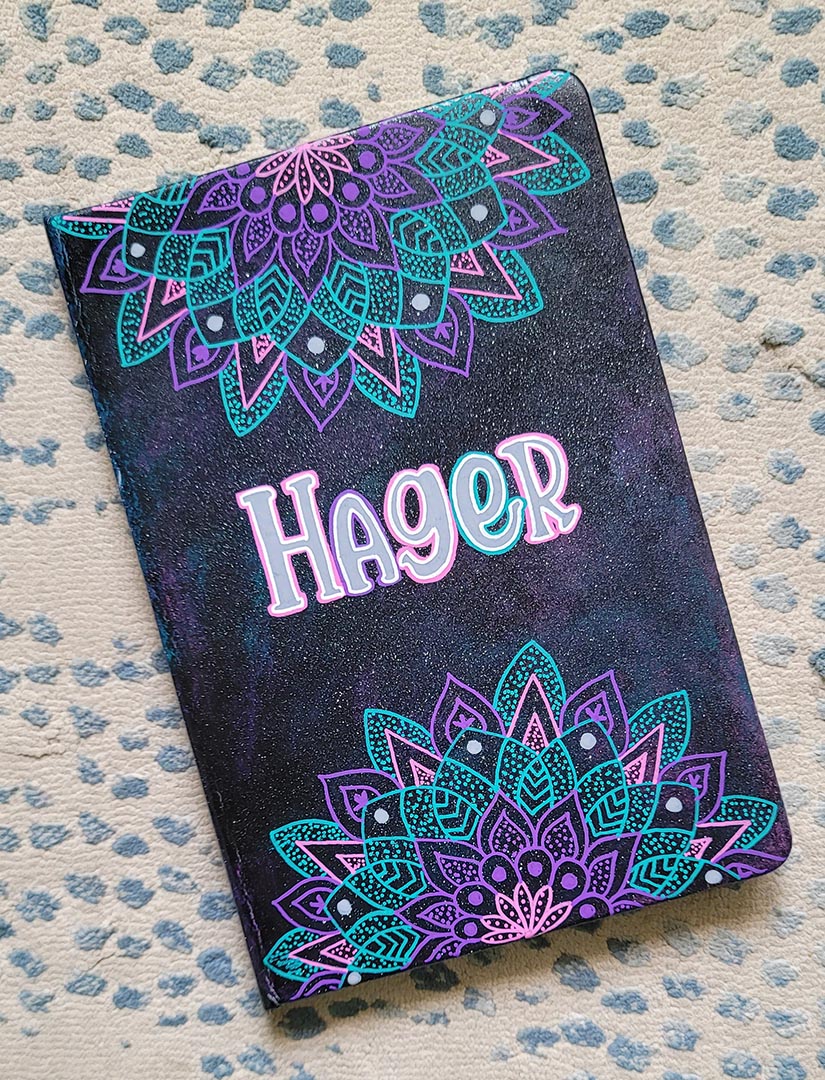 A Black galaxy hand painted noteboo with 2 half mandalas one on top and one at the bottom with the name "Hagar" in the center