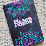 A Black galaxy hand painted noteboo with 2 half mandalas one on top and one at the bottom with the name 