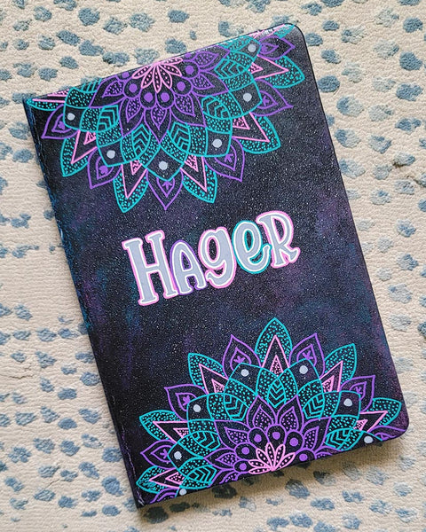 A Black galaxy hand painted noteboo with 2 half mandalas one on top and one at the bottom with the name "Hagar" in the center