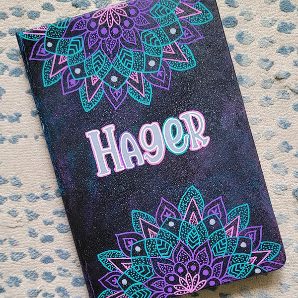 A Black galaxy hand painted noteboo with 2 half mandalas one on top and one at the bottom with the name "Hagar" in the center