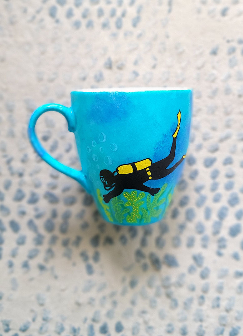 A Blue hand painted mug with a diver painted on it and corals all over the bottom half of the mug