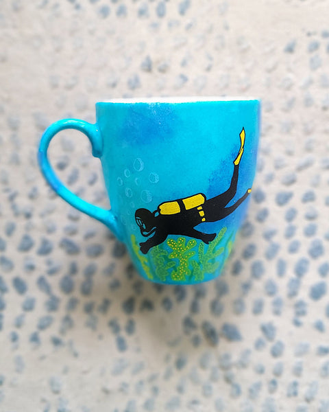 A Blue hand painted mug with a diver painted on it and corals all over the bottom half of the mug