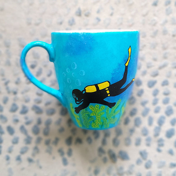 A Blue hand painted mug with a diver painted on it and corals all over the bottom half of the mug