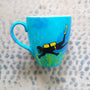 A Blue hand painted mug with a diver painted on it and corals all over the bottom half of the mug