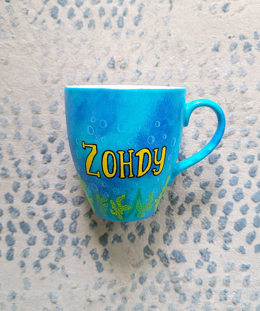 A Blue hand painted mug with the name "Zohdy" and corals all over the bottom half of the mug