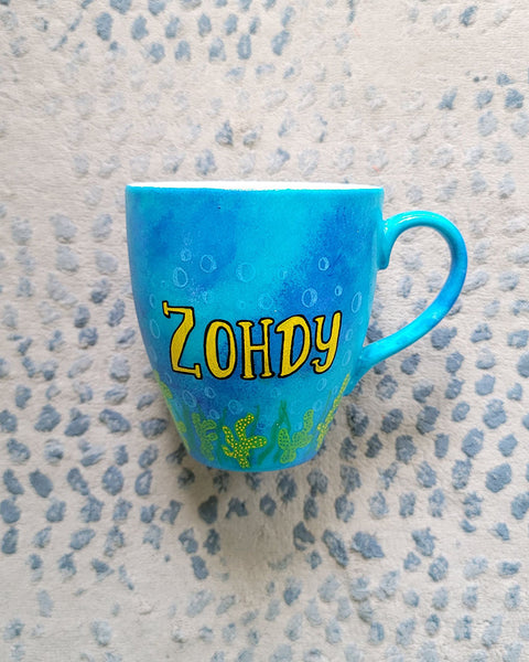 A Blue hand painted mug with the name "Zohdy" and corals all over the bottom half of the mug