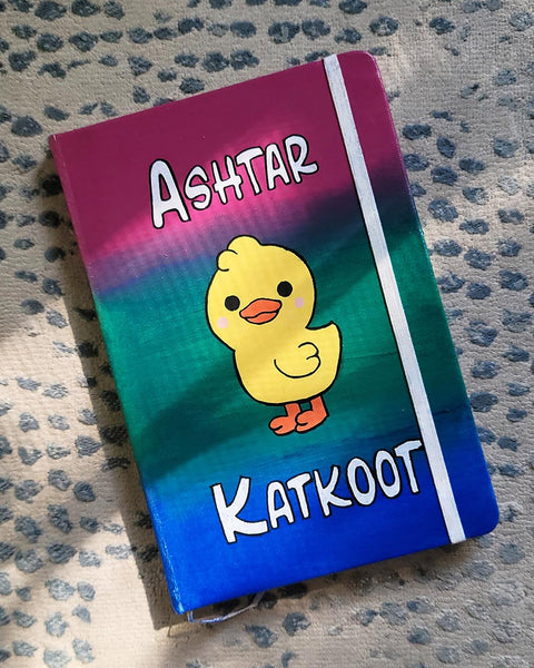 A gradient Hand painted Notebook (pink- green and blue) with the word "Ashtar" on top the the duck painted in the center than the word "Katkoot" below it