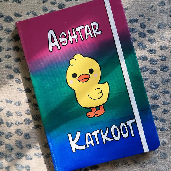 A gradient Hand painted Notebook (pink- green and blue) with the word "Ashtar" on top the the duck painted in the center than the word "Katkoot" below it