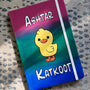 A gradient Hand painted Notebook (pink- green and blue) with the word "Ashtar" on top the the duck painted in the center than the word "Katkoot" below it