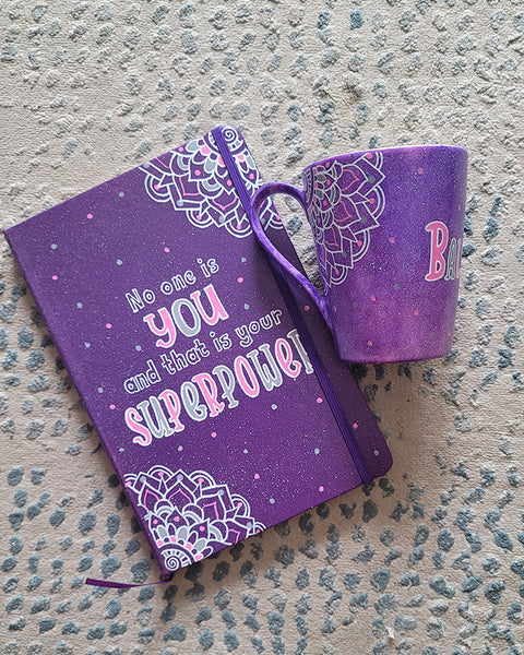 A Gift Set of mug and notebook hand painted in purple with mandalas on 2 opposite corners. The mug has the name balsam and the notebook has the quote "no one is you and that is your superpower"