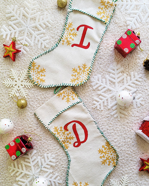 A Picture of two stockings on top of each other ne woht the letter I in red and the other with the Letter D in red, sewn in green thread all around with gold snowflakes in the background