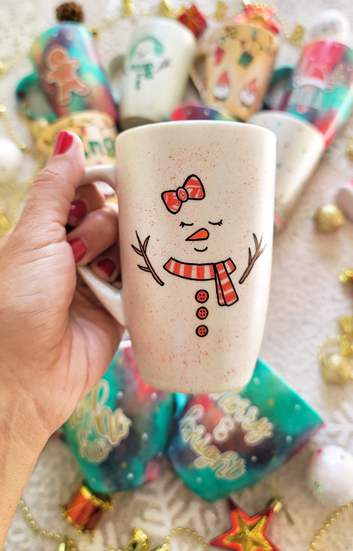 A Pearl white hand painted mug with a red bow drawn on the side and 2 closed eyes and an orange nose and a mouth smiling. wearing a red scarf and 3 buttons drawn below it and 2 tree branches as hands