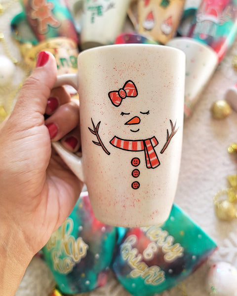 A Pearl white hand painted mug with a red bow drawn on the side and 2 closed eyes and an orange nose and a mouth smiling. wearing a red scarf and 3 buttons drawn below it and 2 tree branches as hands