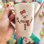 A Pearl white hand painted mug with a red bow drawn on the side and 2 closed eyes and an orange nose and a mouth smiling. wearing a red scarf and 3 buttons drawn below it and 2 tree branches as hands