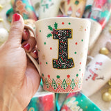 A Pearl white mug with the letter I written filled with doodles in black and colored in green and red. then hz patterns drawn all through the bottom of the mug. with green and red dots, and gold stars scattered all over