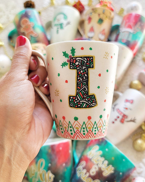 A Pearl white mug with the letter I written filled with doodles in black and colored in green and red. then hz patterns drawn all through the bottom of the mug. with green and red dots, and gold stars scattered all over