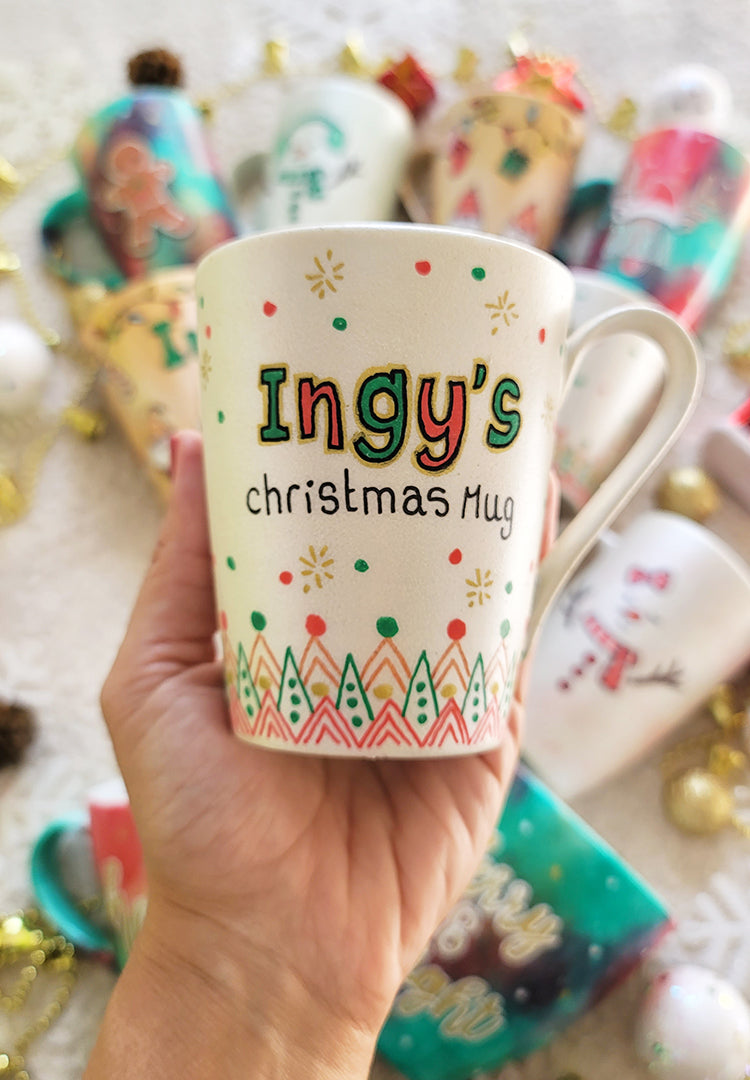 A Pearl white mug with the sentence "Ingy's Christmas Mug". then hz patterns drawn all through the bottom of the mug. with green and red dots, and gold stars scattered all over