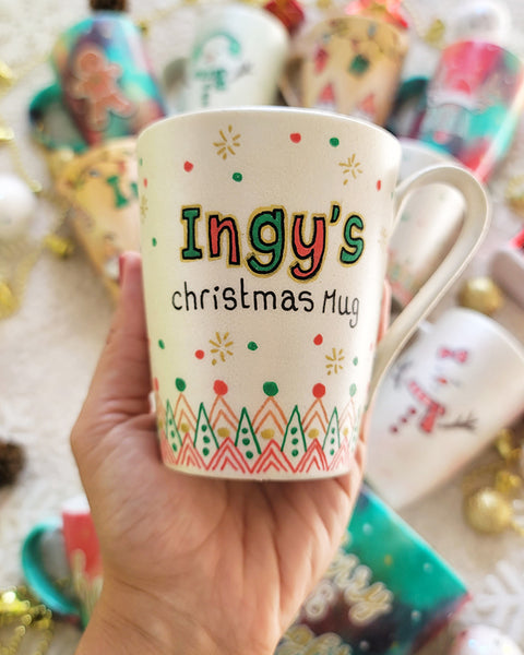 A Pearl white mug with the sentence "Ingy's Christmas Mug". then hz patterns drawn all through the bottom of the mug. with green and red dots, and gold stars scattered all over