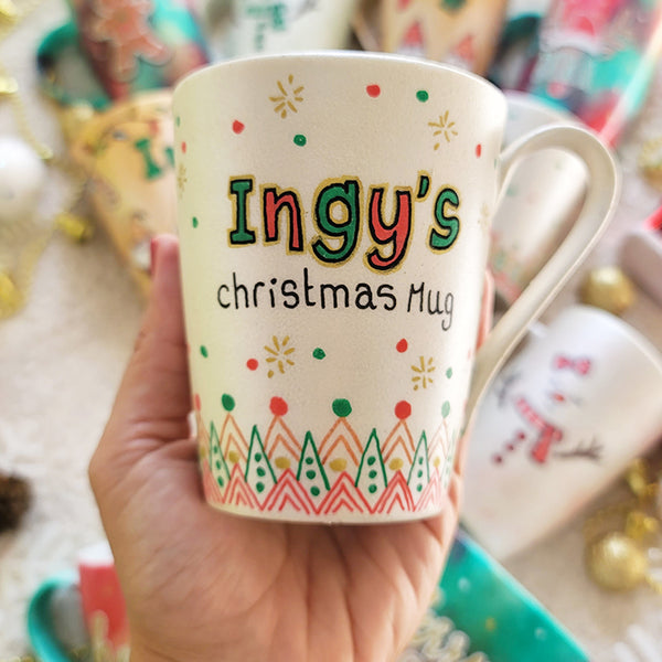 A Pearl white mug with the sentence "Ingy's Christmas Mug". then hz patterns drawn all through the bottom of the mug. with green and red dots, and gold stars scattered all over