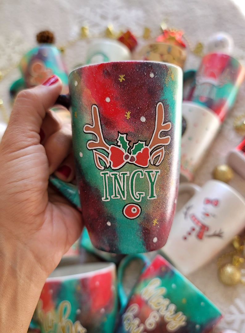 A Christmas galaxy hand painted mug with reindeer antlers with a red bow and reindeer ears, then below it the name "Ingy" and below it the reindeer nose, with white dots and gold stars scattered all over the mug