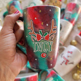 A Christmas galaxy hand painted mug with reindeer antlers with a red bow and reindeer ears, then below it the name 