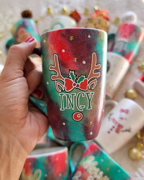 A Christmas galaxy hand painted mug with reindeer antlers with a red bow and reindeer ears, then below it the name "Ingy" and below it the reindeer nose, with white dots and gold stars scattered all over the mug