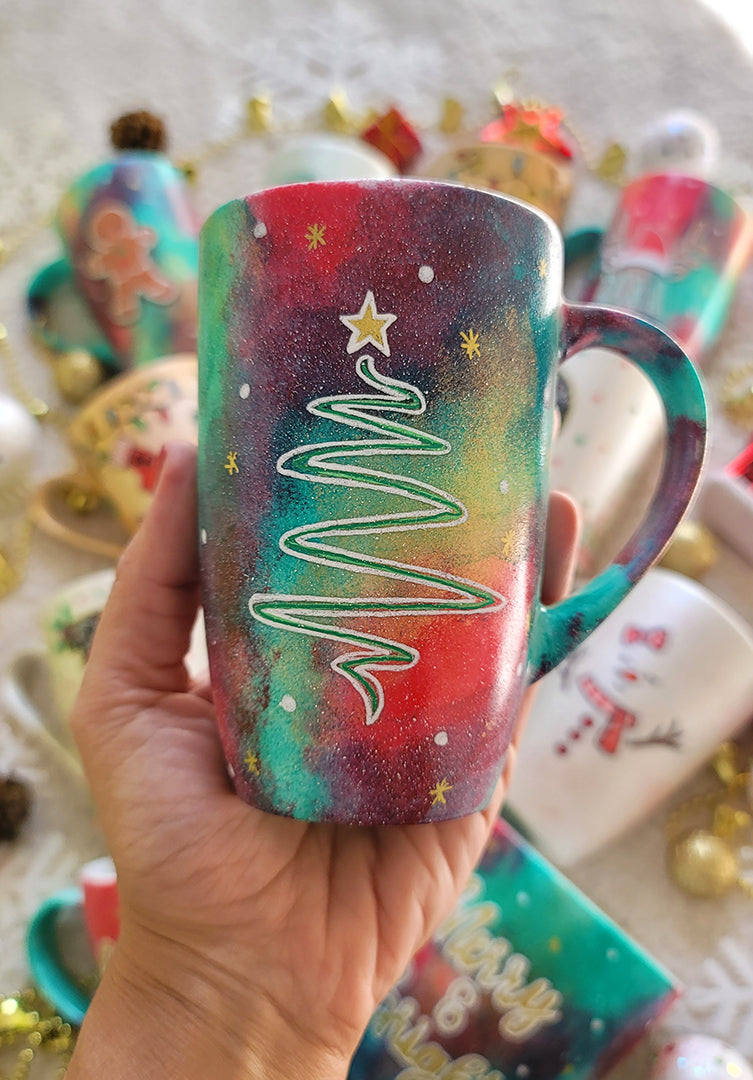 A Christmas galaxy hand painted mug with a christmas linede tree painting in white and colored in green with a gold star on top, with white dots and gold stars scattered all over the mug