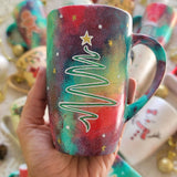 A Christmas galaxy hand painted mug with a christmas linede tree painting in white and colored in green with a gold star on top, with white dots and gold stars scattered all over the mug