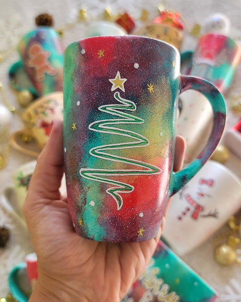 A Christmas galaxy hand painted mug with a christmas linede tree painting in white and colored in green with a gold star on top, with white dots and gold stars scattered all over the mug