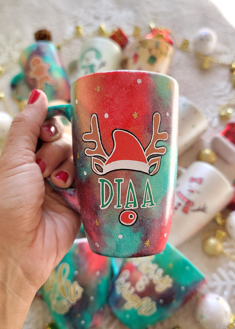 A Christmas galaxy hand painted mug with a reindeer antlers with a santa hat and reindeer ears, then below it the name "Diaa" and below it the reindeer nose, with white dots and gold stars scattered all over the mug