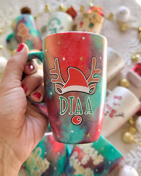 A Christmas galaxy hand painted mug with a reindeer antlers with a santa hat and reindeer ears, then below it the name "Diaa" and below it the reindeer nose, with white dots and gold stars scattered all over the mug