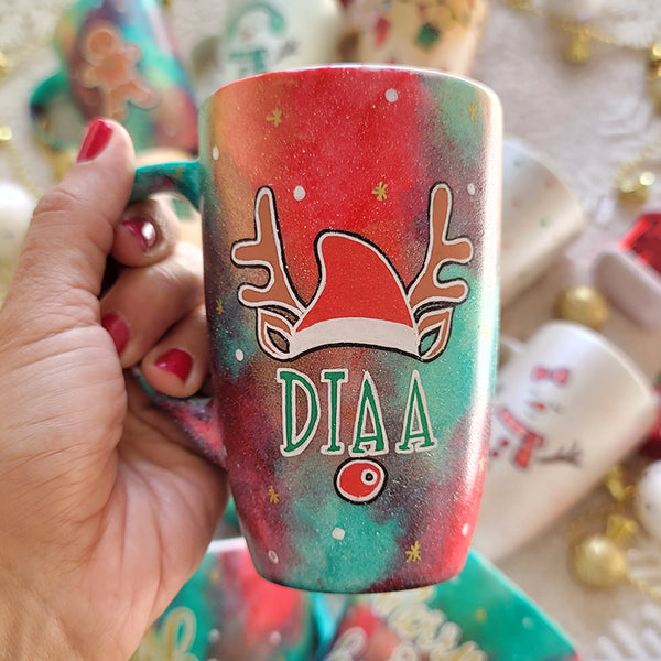 A Christmas galaxy hand painted mug with a reindeer antlers with a santa hat and reindeer ears, then below it the name "Diaa" and below it the reindeer nose, with white dots and gold stars scattered all over the mug