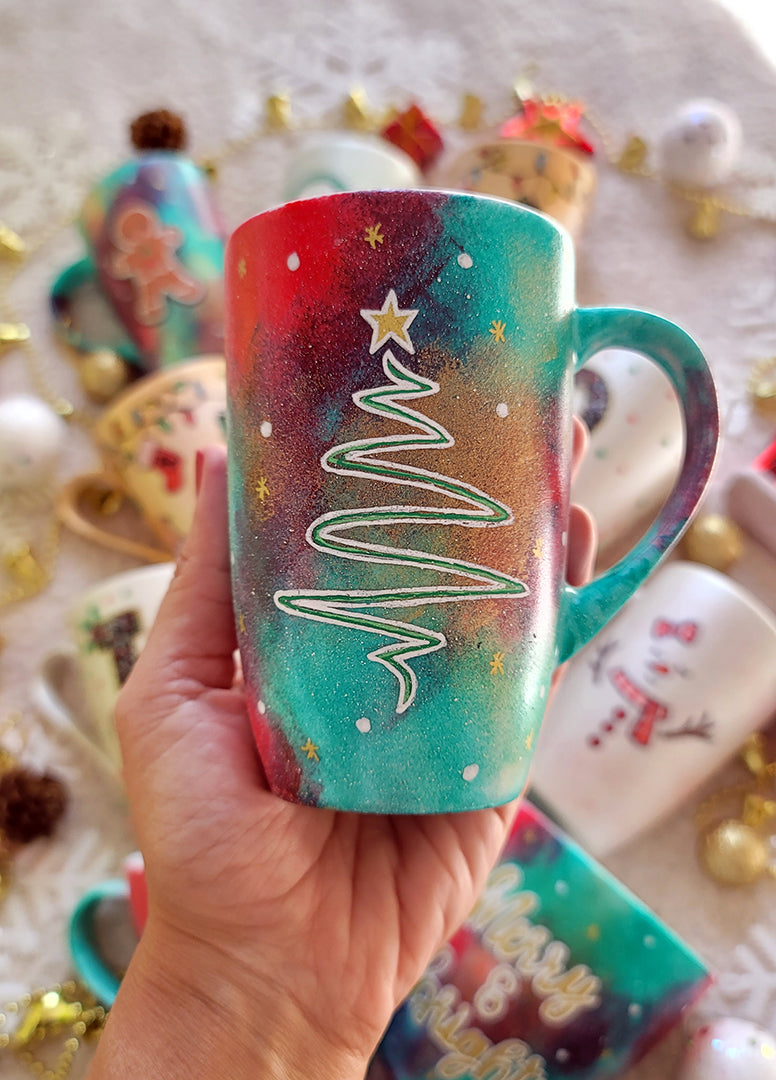 A Christmas galaxy hand painted mug with a christmas linede tree painting in white and colored in green with a gold star on top, with white dots and gold stars scattered all over the mug