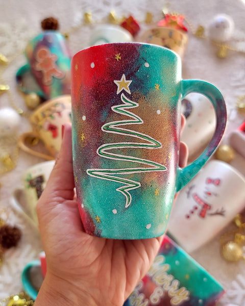 A Christmas galaxy hand painted mug with a christmas linede tree painting in white and colored in green with a gold star on top, with white dots and gold stars scattered all over the mug