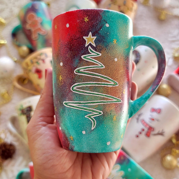 A Christmas galaxy hand painted mug with a christmas linede tree painting in white and colored in green with a gold star on top, with white dots and gold stars scattered all over the mug