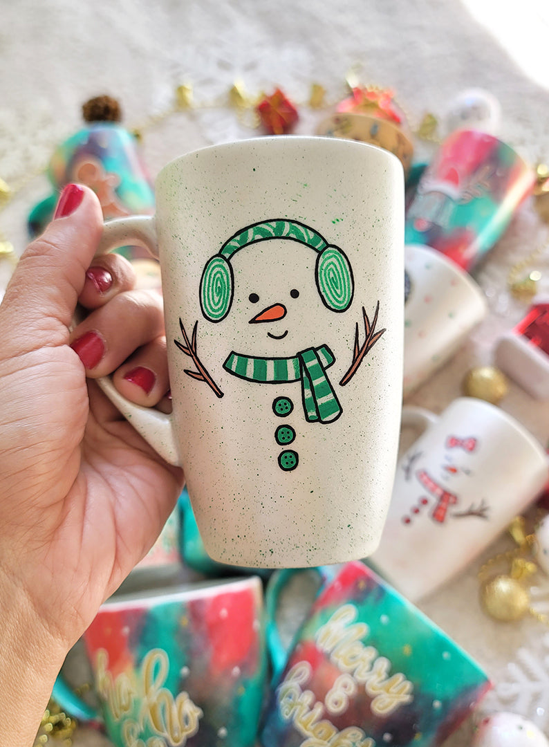 A pearl white hand painted mug with a snow boy painted wearing a green Earmuffs and a green scarf with 3 green buttons in the center and hands made out of tree branches. with an orange nose and a smiling mouth