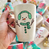 A pearl white hand painted mug with a snow boy painted wearing a green Earmuffs and a green scarf with 3 green buttons in the center and hands made out of tree branches. with an orange nose and a smiling mouth