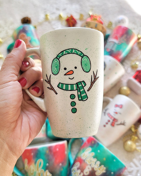 A pearl white hand painted mug with a snow boy painted wearing a green Earmuffs and a green scarf with 3 green buttons in the center and hands made out of tree branches. with an orange nose and a smiling mouth