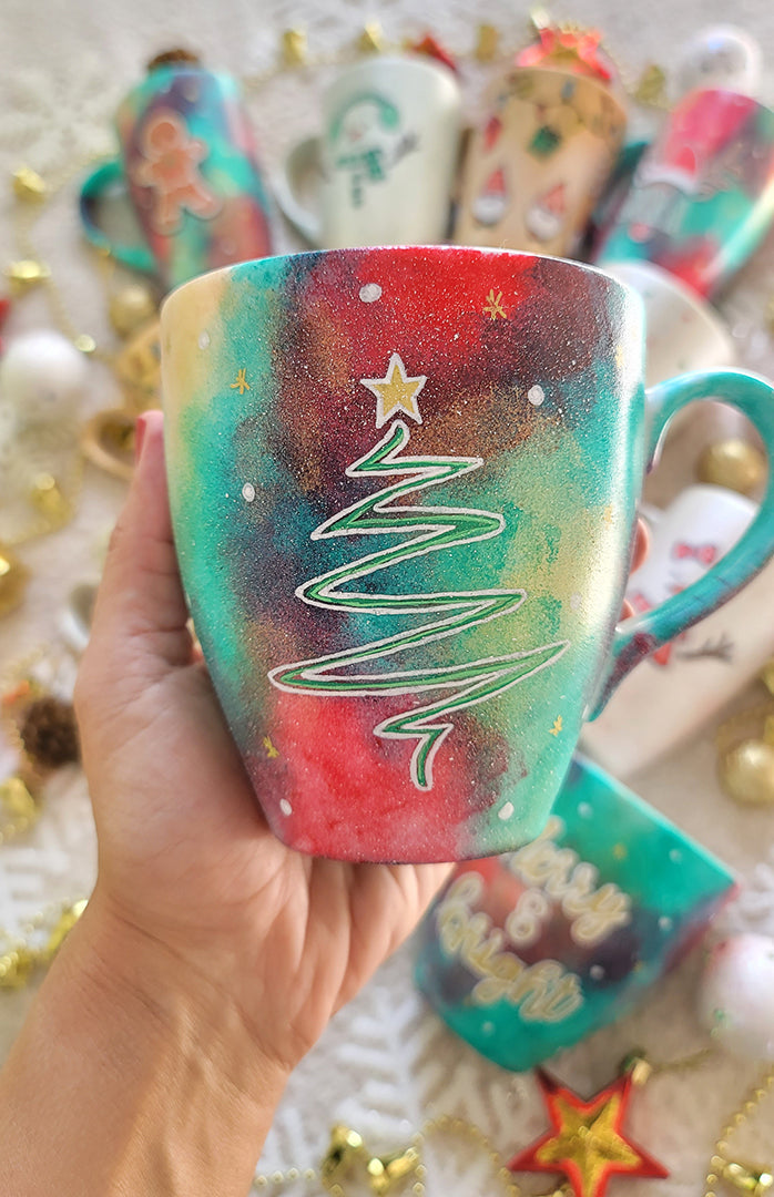 A Christmas galaxy colored hand painted mug with the a line drawing of a christmas tree in white with and green and a gold star on top with white dots and gold stars scattered all over