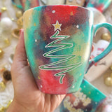 A Christmas galaxy colored hand painted mug with the a line drawing of a christmas tree in white with and green and a gold star on top with white dots and gold stars scattered all over