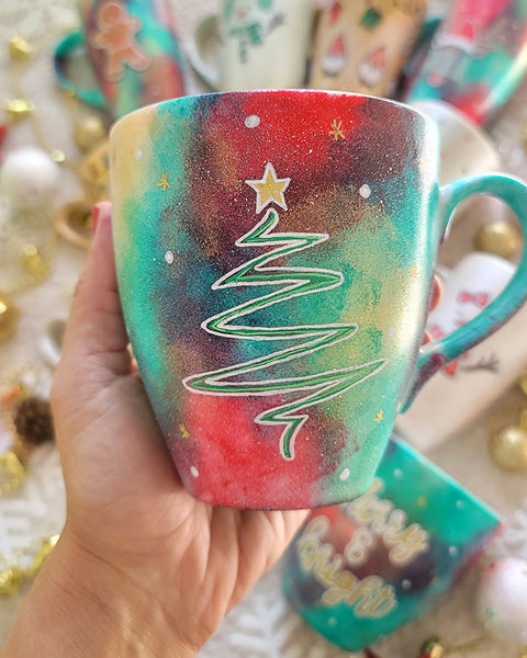 A Christmas galaxy colored hand painted mug with the a line drawing of a christmas tree in white with and green and a gold star on top with white dots and gold stars scattered all over