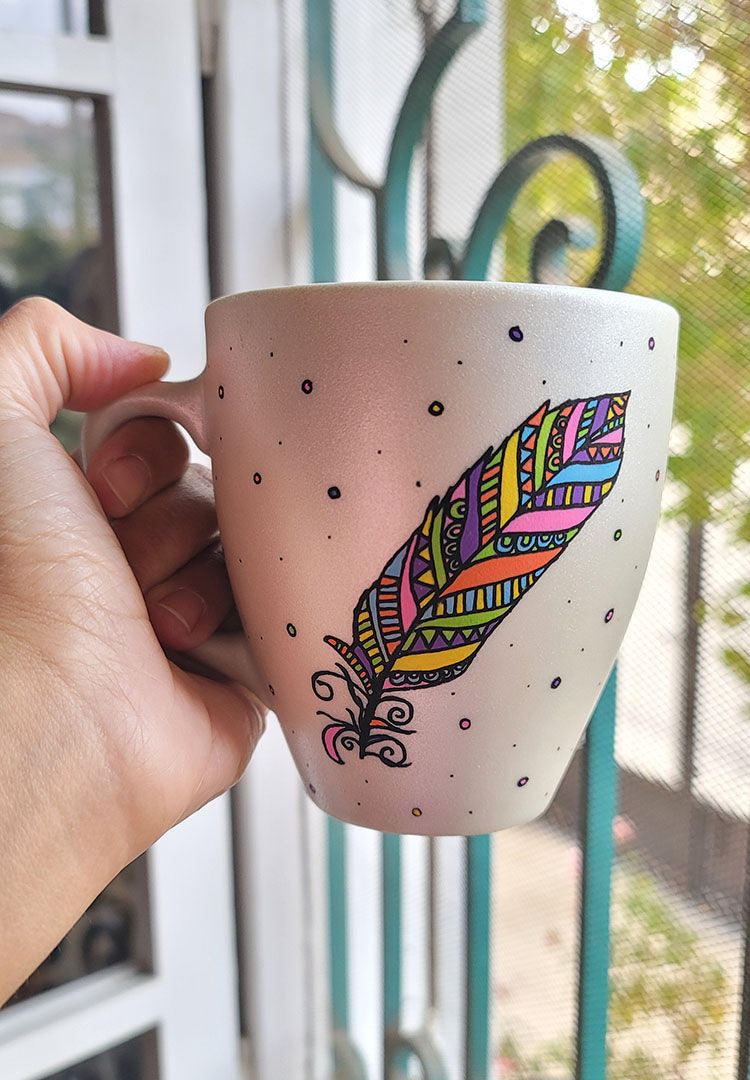 A Pearl white hand painted mug with a colorful feather outlined in black and filled with paterns in colors of orange, yellow, pink, purple, green and blue