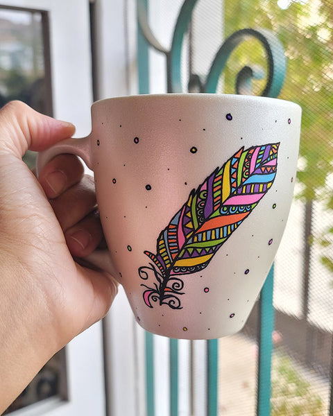 A Pearl white hand painted mug with a colorful feather outlined in black and filled with paterns in colors of orange, yellow, pink, purple, green and blue