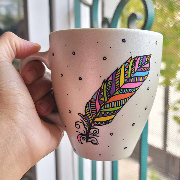 A Pearl white hand painted mug with a colorful feather outlined in black and filled with paterns in colors of orange, yellow, pink, purple, green and blue