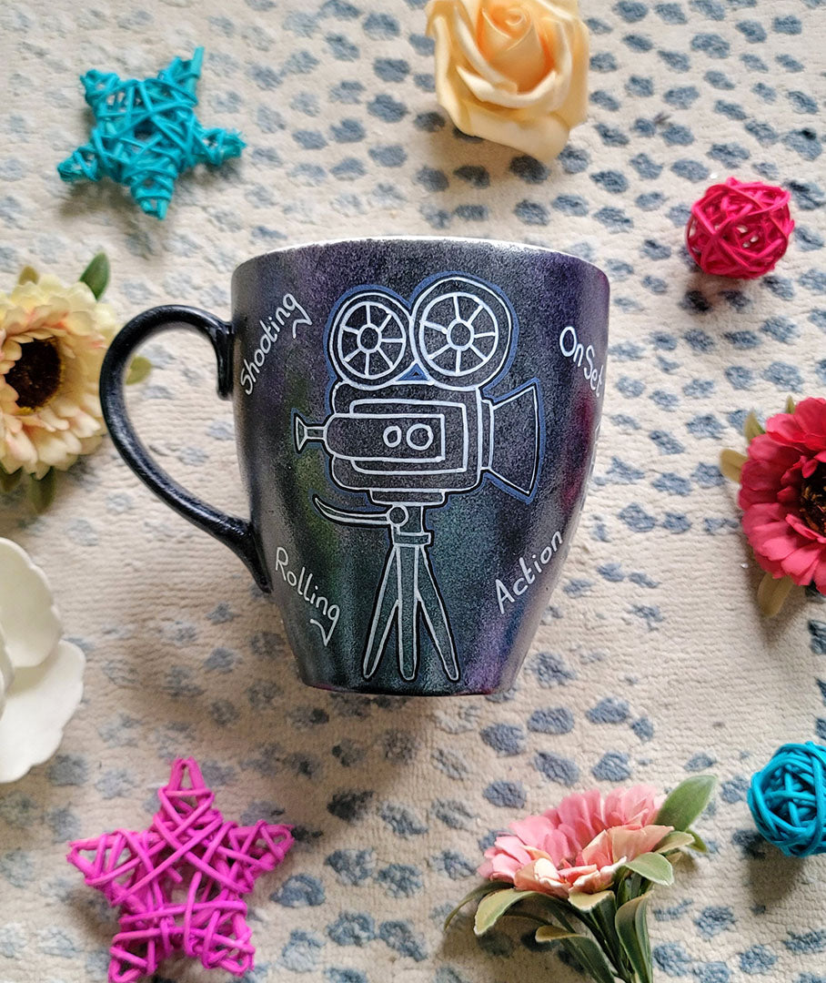 A Dark sky mug with a movie camera painted on it and words "Rolling. on set , Shooting, action" scattered all over